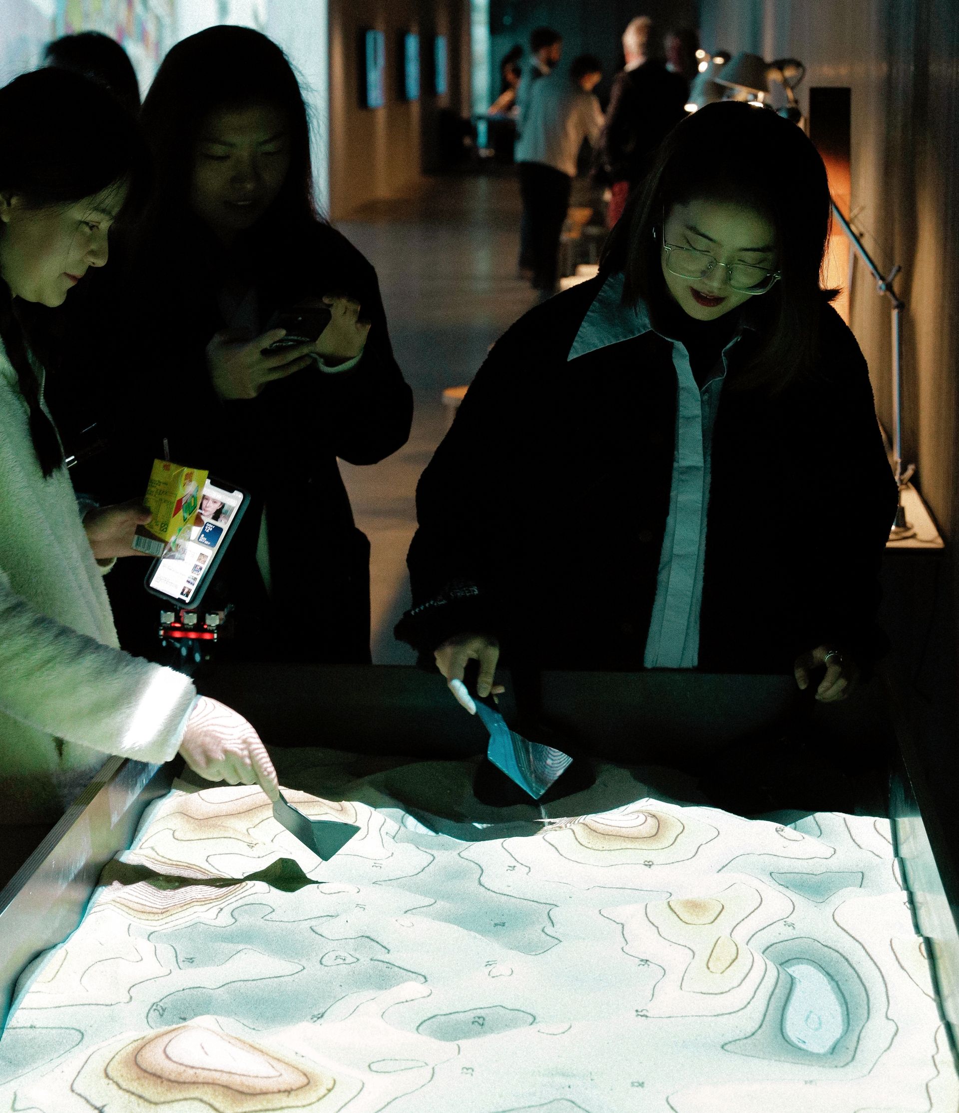 Two people use small sculptural tools to shape sand contained in a box with an overhead projection displaying contour lines and elevations.